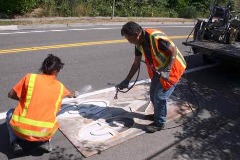Brian Tanaka sprays pain onto the surface of Lakeview Drive through a bike lane stencil while co-worker Debra Eastburn throws down some powder to give it texture. This was just one of 560 bike lane symbols to be repainted.